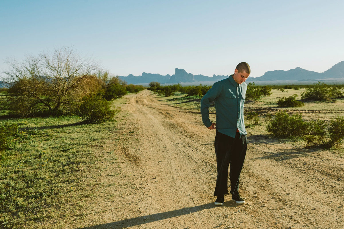 Man in a long-sleeve button-up shirt standing on a desert road with mountains behind